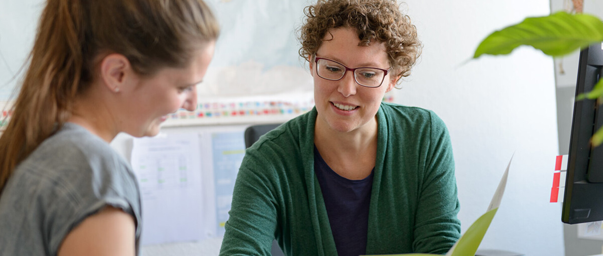 Zwei Frauen sitzen an einem Schreibtisch und führen ein Gespräch. Eine Frau mit lockigen Haaren und Brille zeigt der anderen etwas. Im Hintergrund sind Büromaterialien und eine Pflanze sichtbar.