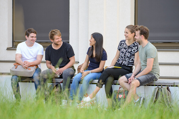 Eine Gruppe Studierender sitzt nebeneinander auf einer Bank im Außenbereich von B6. 
