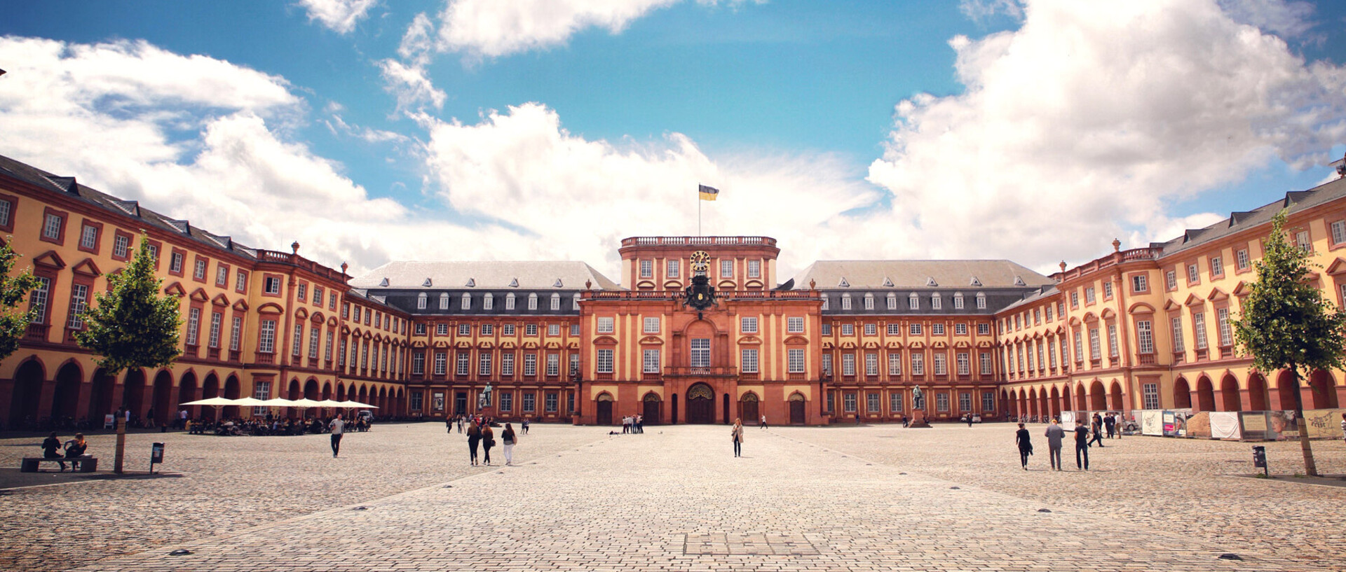 Das Barock-Schloss und der Ehrenhof der Universität Mannheim unter strahlend blauem Himmel. Das Schloss ist von unzähligen Fenstern, rotem Sandstein und einer gelben Fassade geprägt.